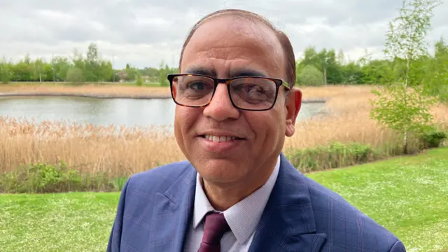Mohammad Yasin standing on a grassy bank next to a lake. There are reeds at the edge of the lake. He is wearing a blue jacket, lilac shirt and mauve tie.
