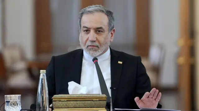 Seyed Abbas Araghchi sits at a table in front of a microphone with a box of tissues, bottle and glass of water beside him
