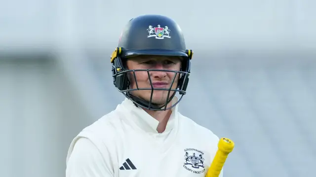 Miles Hammond wearing a Gloucestershire navy helmet with his yellow bat handle under his left arm