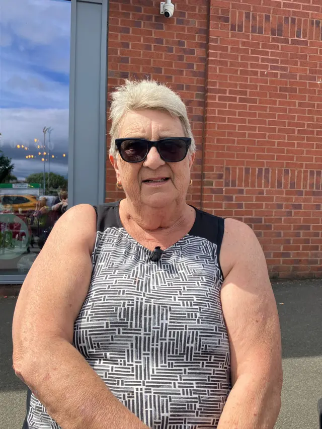 A woman wearing a black and white top and sunglasses stands outside, with a brick wall behind her.