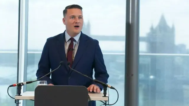 Wes Streeting, a white man with short dark hair, stands behind a podium speaking into microphones. Behind him is a glass window with the outline of tower bridge in London visible through frosted glass.