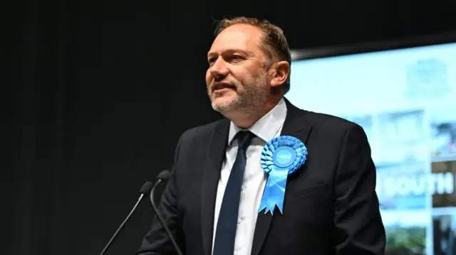 Douglas Lumsden with a rosette and standing at a lectern