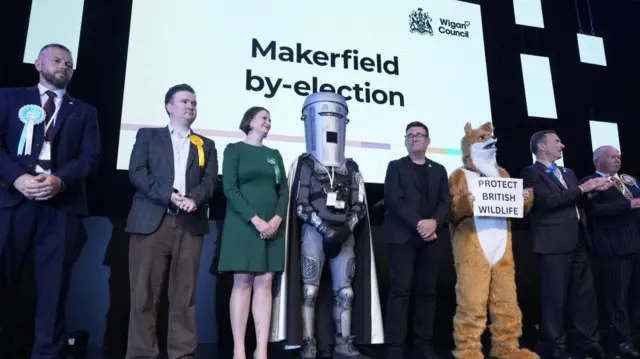 Andy Burnham stands at the centre of the group of candidates in Makerfield. Reform's Robert Kenyon and Count Binface are among others