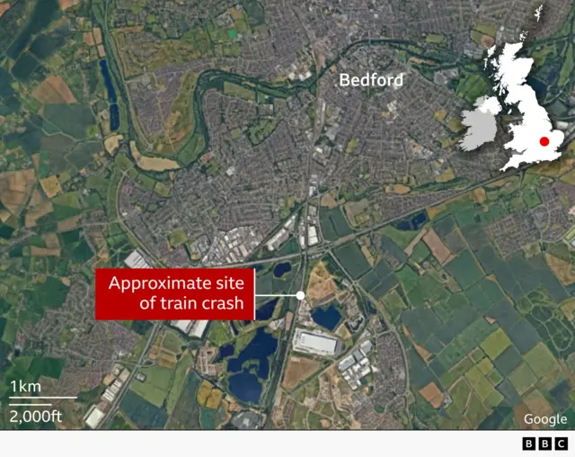 Satellite map of Bedford and surrounding countryside in England. The town centre is labelled “Bedford” near a winding river running across the upper half of the image. A red box and pointer mark the “approximate site of train crash” to the south of the town, near to an industrial site. An inset map of the UK highlights the location in Bedfordshire with a red dot.
