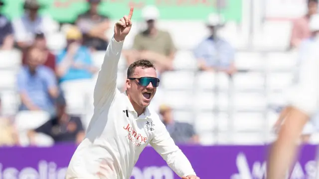 Liam Patterson-White wearing sunglasses celebrates taking a wicket with his right index finger pointing to the sky