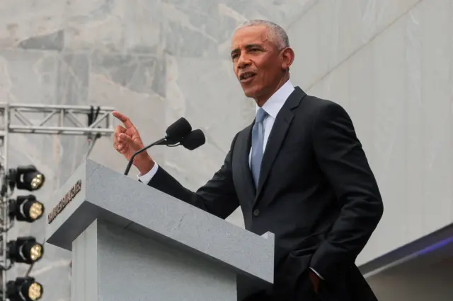 Barack Obama stands at a lectern in a black suit and blue tie while pointing his right hand towards the crowd during a speech in Illinois