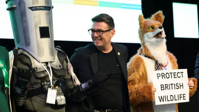 Andy Burnham smiles as he looks at his fellow candidate Count Binface. Another candidate dressed as a fox with a sign saying 'protect British wildlife' is on Burnham's other side