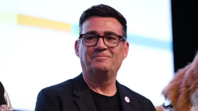 Andy Burnham smiles while standing on a stage. He is wearing a dark-coloured suit.