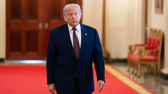 US President Donald Trump in a blue suit and red tie, walking on a red carpet inside a building.