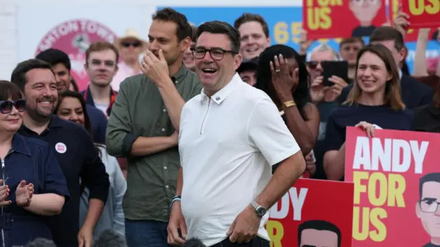 Andy Burnham in a white top smiling as people crowd round him smiling, signs behind him say Andy for Us.