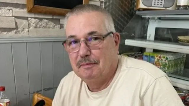 Keith Crossland, an older white man with short hair and glasses in a cream t-shirt sat in a cafe.