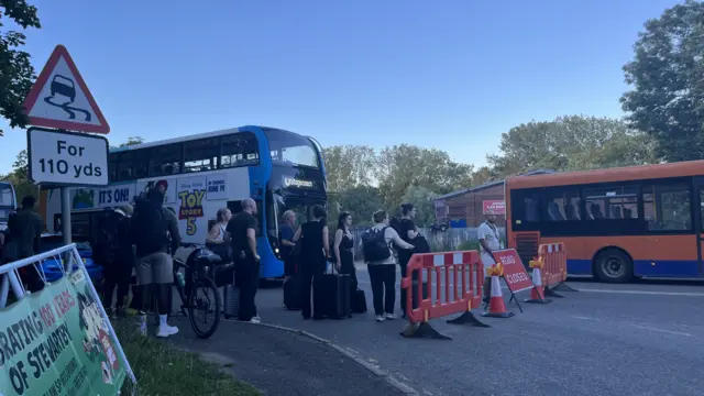 People wait in a queue to get on to several buses parked on a road.