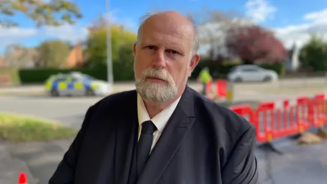 Mayor of Bedford Tom Wootton, who has a bald head and grey beard, looks into the camera while wearing a black suit, white shirt and black tie. He is standing outside next to some orange barriers, with a police car and officer in the background