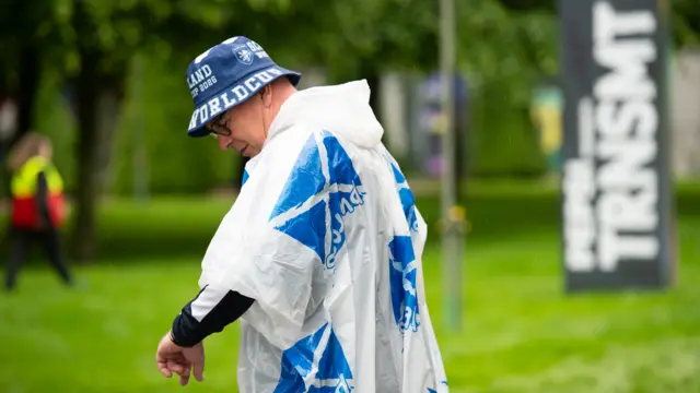 Fans arrive at TRNSMT festival in the rain. A man wearing a poncho and a blue Scotland beannie hat is looking at his watch,