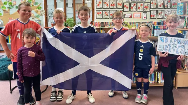 Seven pupils stand together in a classroom holding a Scotland flag.