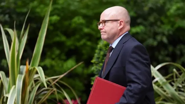 Attorney general Richard Hermer is pictured walking against a background of trees, wearing a suit and carrying a red folder