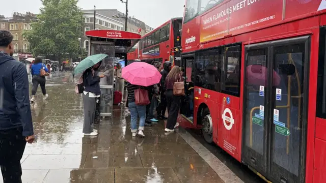 A crowd of people are grouped close to the front door of a bus as they board from a bus stop in the rain
