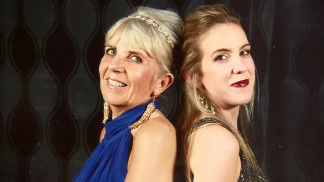 Two women are standing back to back. One has blond hair and a fringe. The other has long brown hair.
