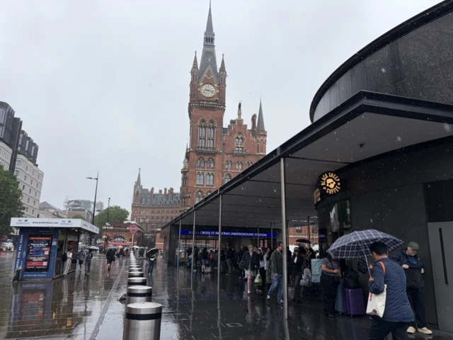 A view from outside Kings Cross as people shelter under a covered walkway near the entrance and the station's clock tower is shown in the background