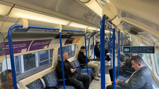 Interior of a London Underground train carriage showing seated passengers, blue handrails, and advertising panels above the windows.