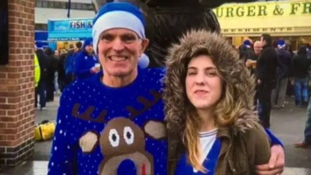 A man and woman are smiling at the camera. He is wearing a a blue Christmas jumper and hat. The woman is also wearing a blue jumper and a coat with a fluffy hoot which is up over her head.
