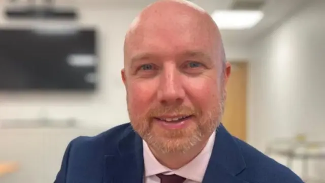 A head shot of a bald man smiling. He is wearing a blue suit jacket with a white shirt and a maroon tie.