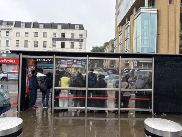 A large queue of people are photographed from behind while inside a bus stop outside Kings Cross