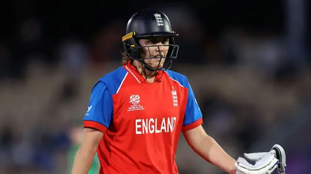 Nat Sciver-Brunt, wearing a red England shirt with blue shoulders and a dark blue helmet, walks off the field