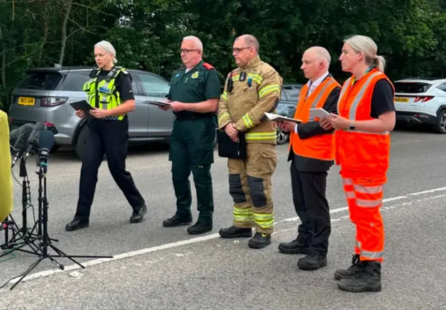 Three men and two women stood on a road speaking into microphones, two are wearing high vis outfits, one is in a police officer's outfit, one is in paramedic gear and the other is a firefighter.