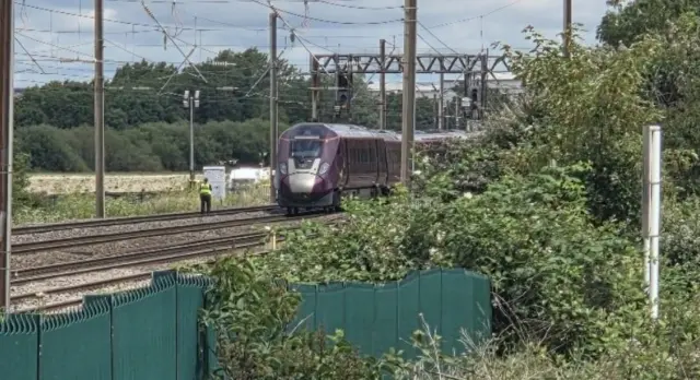 A view of the front of a train stopped on the tracks, shrubbery around and a person in high vis stood next to the train.