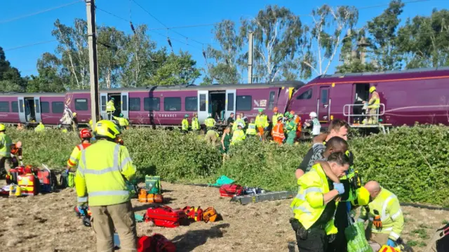 Emergency service personnel tend to passengers, while two collided trains are visible in the background.