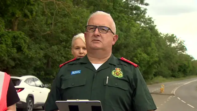 Paul Gates, Strategic Commander for the East of England Ambulance Service, in green uniform, stands in the middle of a closed off road addressing the press. Behind him is a blonde woman