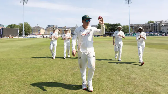 Liam Patterson-White wearing a green cap and sunglasses leads his team off while raising the ball in his left hand to acknowledge the applause of spectators