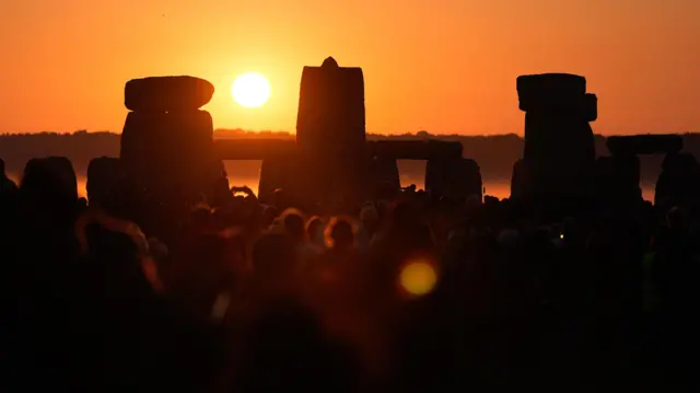The sun peeks over the top of the stone circle at Stonehenge as a crowd watches on.