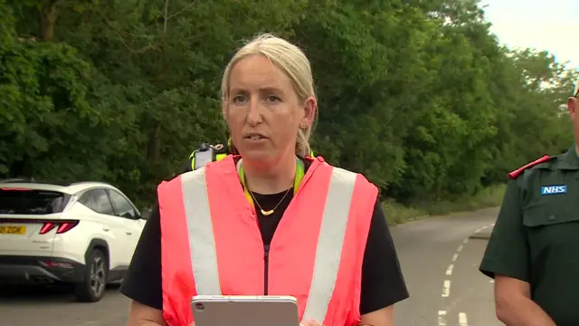 Regional Managing Director for Network Rail East Ellie Burrows in a pink hi-vi jacket and short-sleeved black shirt addresses the media from the middle of a closed off road