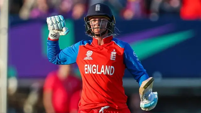 Danni Wyatt-Hodge, wearing a red England shirt with long blue sleeves and a dark blue helmet, raises a clenched right fist and holds her bat in her left hand