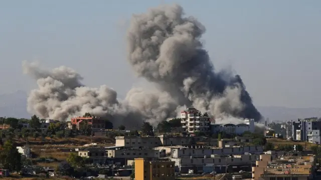 Smoke billows from southern Lebanon with hills with buildings and trees in the foreground and two huge plumes of smoke