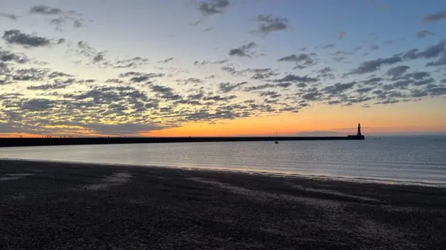 A view of a beach and the sea with a light house in the distance. The sun is rising on the horizon.