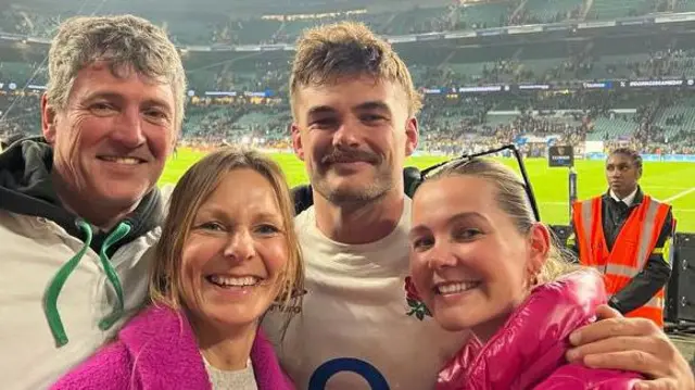 Four people are standing in front of a pitch where a team has played rugby. They are all smiling.