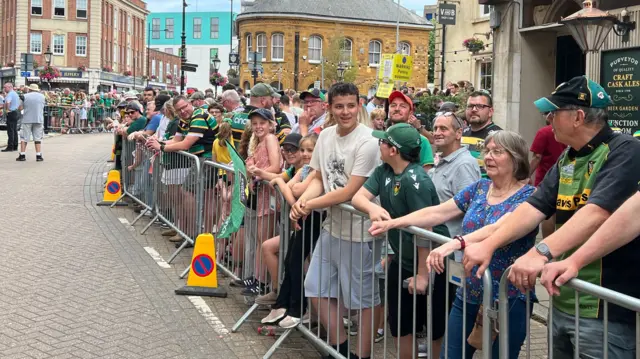Hundreds of people are standing behind a barrier and are ready for a parade to take place. Several of the are wearing Northampton Saints rugby shirts.