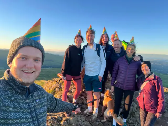A group stood on top of a mountain peak, wearing party hats.