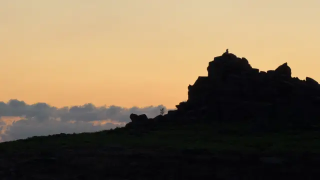 A view of rocks where a person is sitting at the top watching the sunrise.