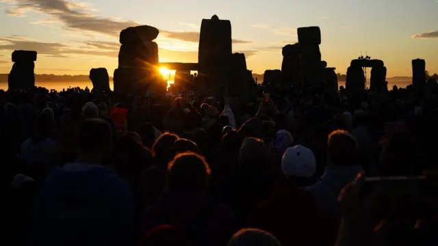 The Sun rises between the stones at Stonehenge with people's silhouettes in the foreground.