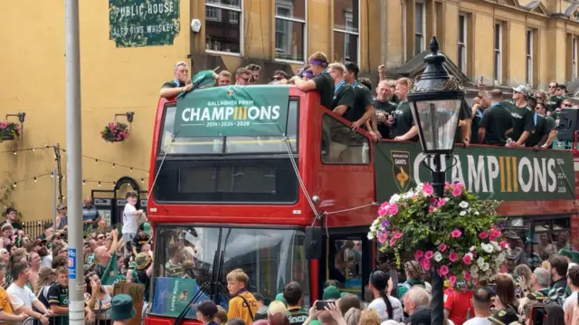A double decker red bus is heading through a town centre. Rugby players are standing at the top and are surrounded by fans watching.