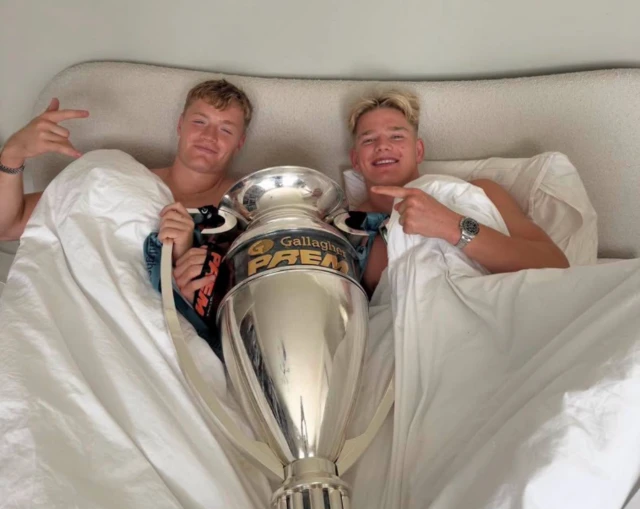 Two of Northampton Saints players are in a bed which has white bedding. In between them is a silver trophy which Northampton Saints won.
