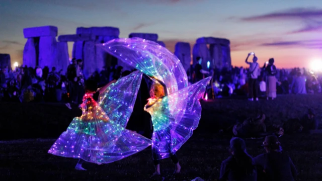 Two people wearing dresses lit up in different colours are dancing by Stonehenge.