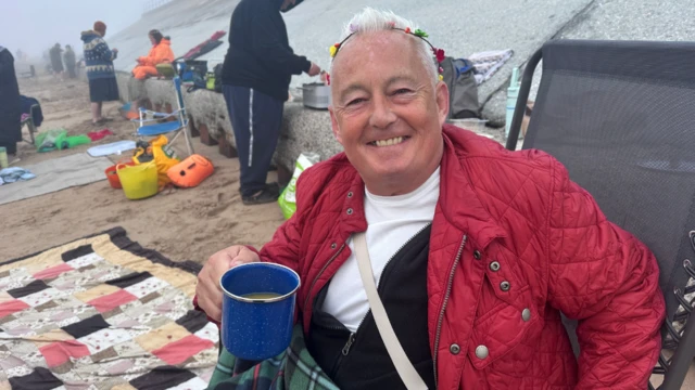 A man is sitting in a camping chair on the beach holding a cup of tea.
