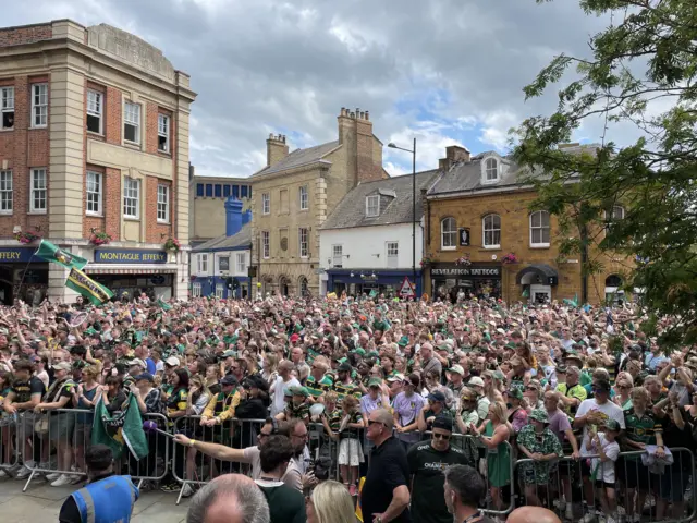 Hundreds of people are standing in a town centre and are behind a metal railing. Most of them are wearing Northampton Saints T-shirts and hats.