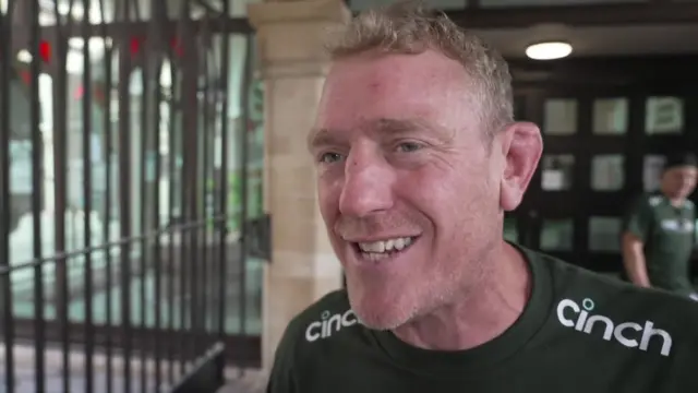 Sam Vesty has short brown hair. He is wearing a green T-shirt which has branding on it. He is standing in the Guildhall.