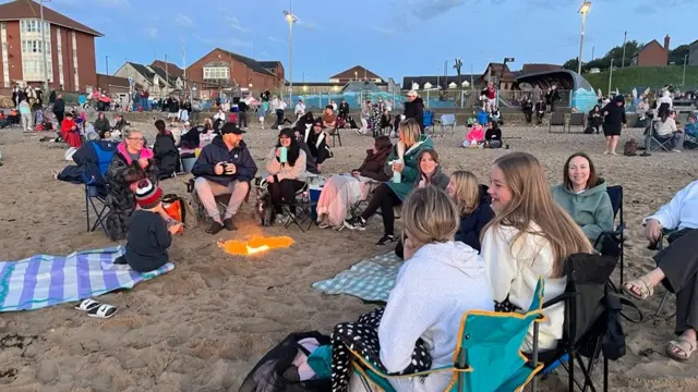 People are sitting on camping chairs around a fire on the beach.
